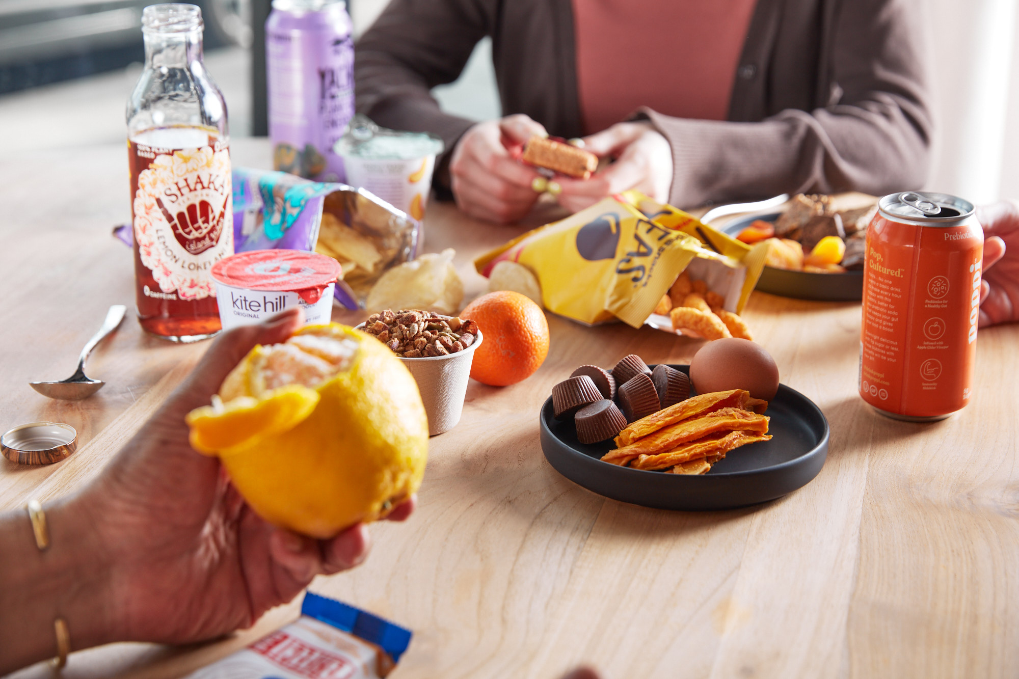 A close-up tabletop snack spread with bottled tea, a Poppi soda, yogurt, chips, an orange, chocolate, dried mango, and trail mix