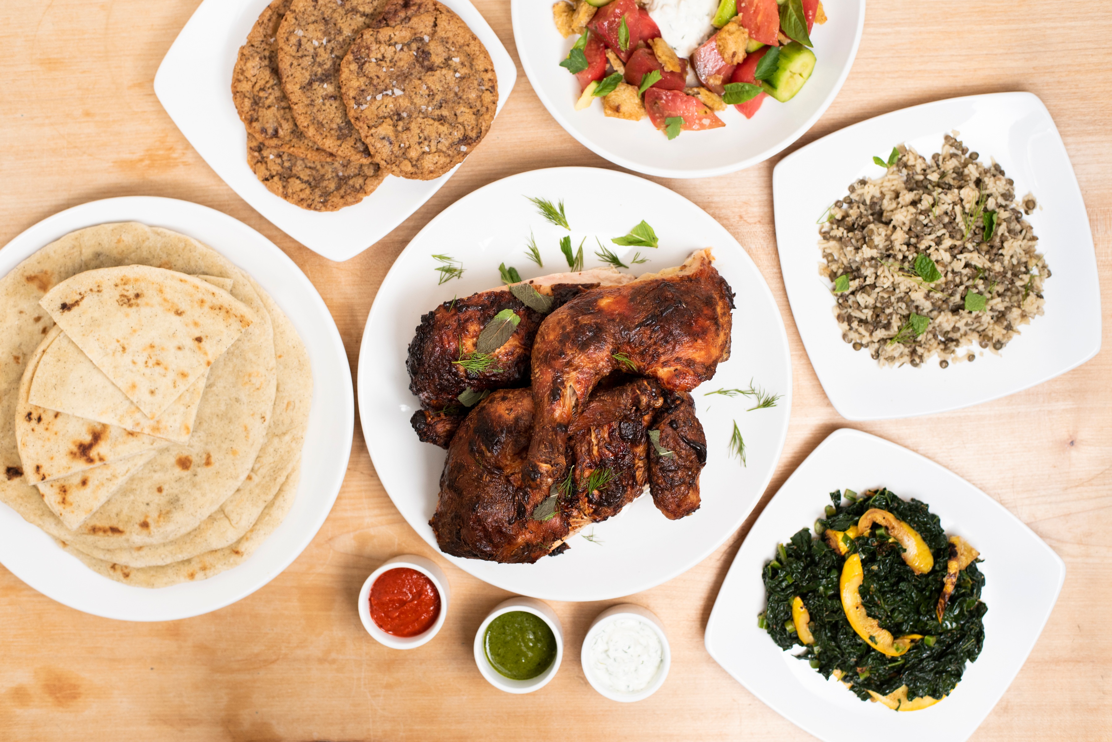An overhead spread of Just Fare dishes on a wooden table with charred tandoori-style roasted chicken, flatbread, a tomato-cucumber salad, lentil-and-rice pilaf, sautéed greens, cookies, and dipping sauces