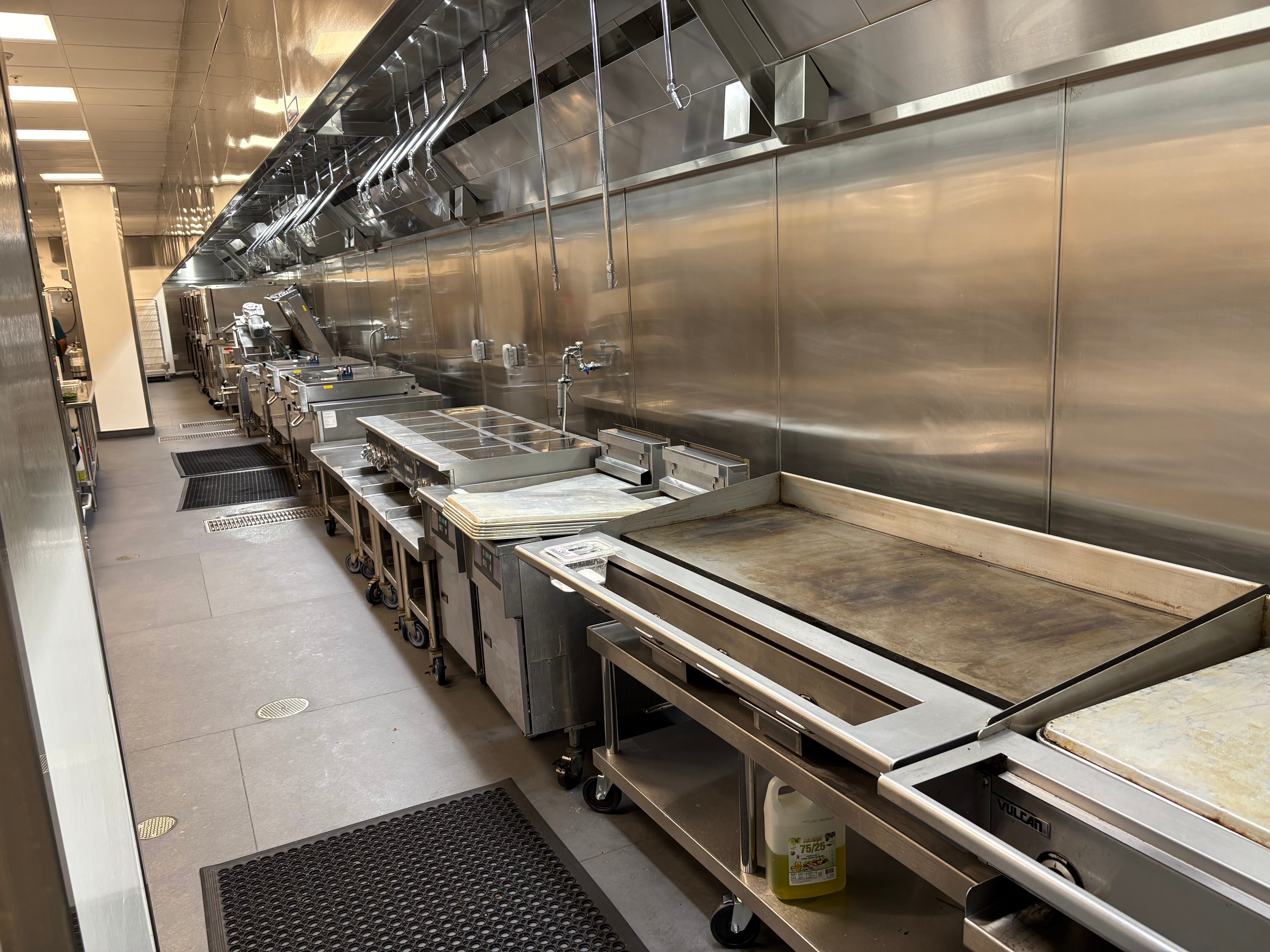 Stainless steel commercial kitchen line with griddle, fryers, and prep tables under an exhaust hood