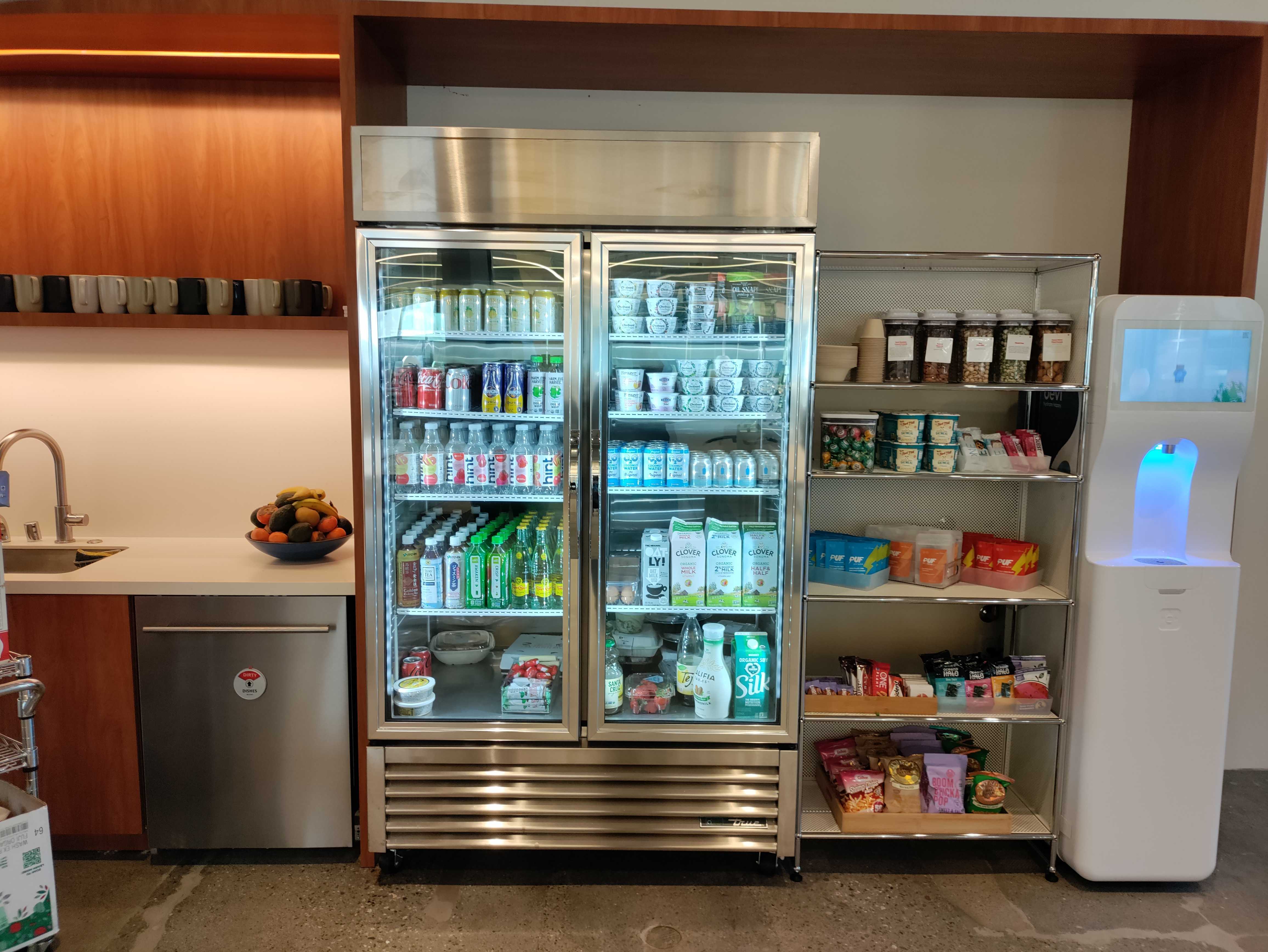 Office micro-kitchen with a stocked glass-door beverage fridge, chrome shelving of bulk snacks and yogurts, a fresh fruit bowl, and a touchscreen water dispenser