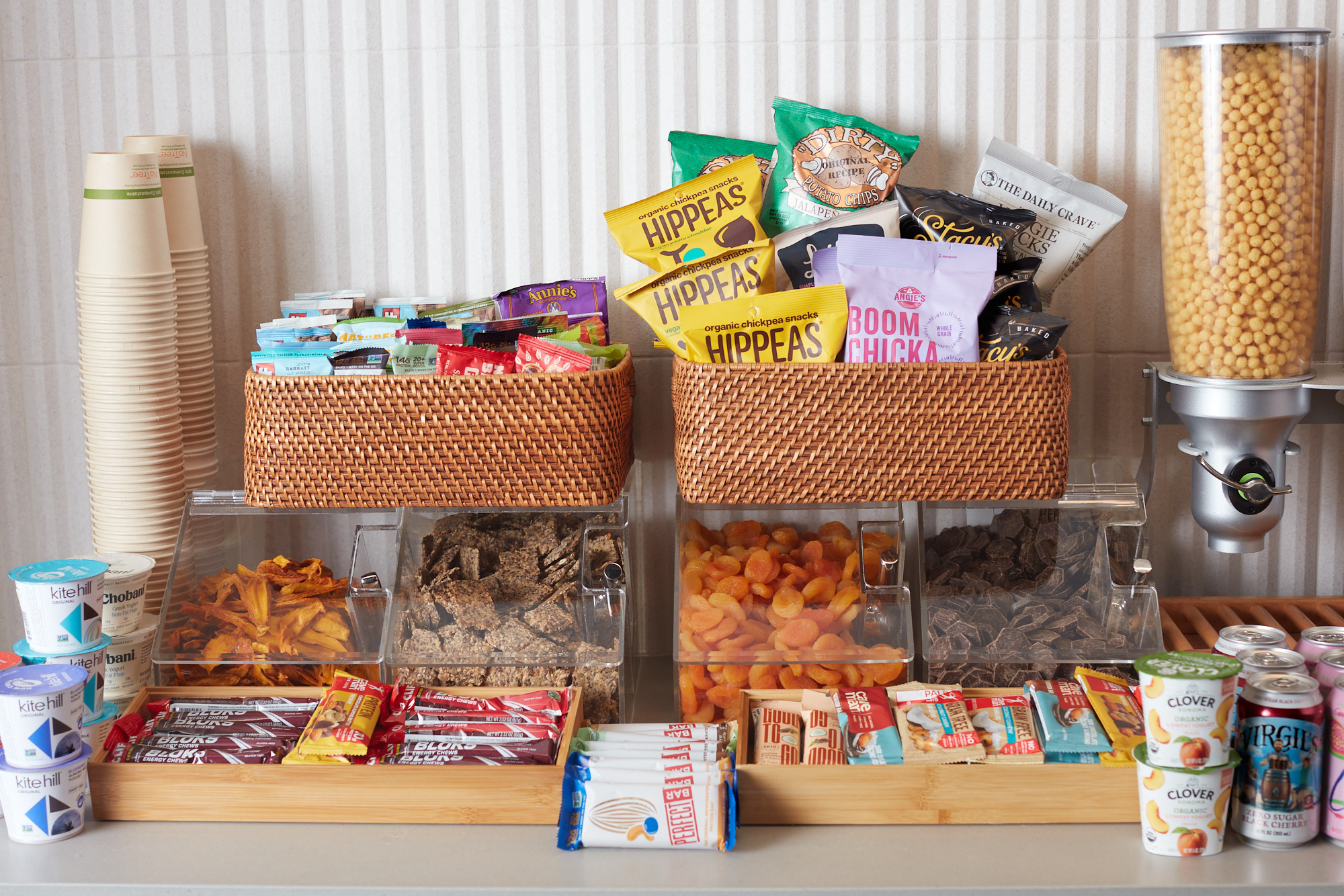 Fully stocked office snack station with baskets of chips, bins of dried fruit, energy bars, yogurts, and a cereal dispenser